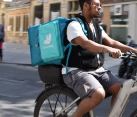 Deniz · 09/05/2026 · 6000 x 4000 Cette photographie montre un livreur à vélo, sujet principal, portant un sac teal de livraison. Le cadrage est serré, mettant en valeur son visage et son activité.La composition est dynamique, avec une route en arrière-plan légèrement floue. La lumière est naturelle et claire, suggérant un jour ensoleillé. Les couleurs dominantes sont les tons neutres (gris de la route, beige des bâtiments) et le bleu vif du sac, apportant une touche moderne.Le style est documentaire et réaliste, capturant une scène urbaine du quotidien. L’ambiance est active et professionnelle.