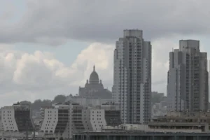 Pierre · 03/05/2026 · 2523 x 1678 Cette photographie montre une ville avec des gratte-ciels modernes en premier plan. Le sujet principal est un ensemble architectural contrasté : des bâtiments contemporains et une cathédrale historique en arrière-plan, partiellement visible.La composition est équilibrée, avec un cadrage large capturant une partie du ciel nuageux. La lumière est diffuse, atténuée par les nuages, créant une ambiance sobre. Les couleurs dominantes sont les tons gris et beiges, typiques d’un jour couvert. Le style est réaliste et documentaire, évoquant une atmosphère urbaine à la fois dynamique et mélancolique.