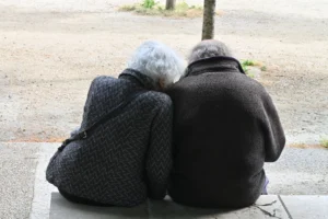 Pierre · 03/05/2026 · 6048 x 4024 Cette photographie montre deux personnes âgées assises dos à nous, enlacées sur un banc. Le **sujet principal** est leur complicité silencieuse et leur proximité physique.La **composition** est simple et centrée, avec un **cadrage serré** qui isole les deux silhouettes. La **lumière**, douce et naturelle, semble tamisée, créant une ambiance calme et mélancolique. Les **couleurs dominantes** sont neutres, avec des tons gris et beiges, renforçant une impression de sobriété et de sérénité.Le **style** est documentaire et intimiste, évoquant une **ambiance** de tendresse et de nostalgie.