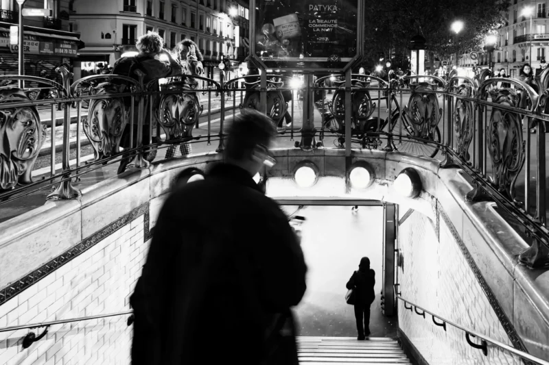 Bao · 05/11/2025 · 6700 x 4466 Cette photographie en noir et blanc capture une scène urbaine nocturne. Le sujet principal est un homme en manteau, vu de dos, descendant un escalier. La composition joue sur les lignes et les niveaux, avec des escaliers et des passerelles métalliques.Le cadrage est légèrement incliné, créant une perspective dynamique. La lumière est douce et tamisée, éclairant faiblement les zones centrales et laissant les bords dans l’ombre. Les couleurs dominantes sont les nuances de gris, renforçant l’atmosphère nocturne et mystérieuse. Le style est documentaire et réaliste, évoquant une ambiance calme et mélancolique.