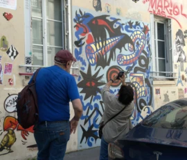 Bao · 5317 x 4016 Cette photographie montre deux personnes observant un mur couvert de graffitis colorés. Le sujet principal est l’art urbain, avec des motifs variés et dynamiques, incluant des silhouettes et des éléments stylisés.La composition est équilibrée, avec les personnages en premier plan et le mur en arrière-plan. Le cadrage est serré, mettant en valeur les détails des fresques. La lumière est naturelle, légèrement tamisée, créant des contrastes doux.Les couleurs dominantes sont vives et contrastées : bleus, rouges, noirs et blancs. Le style est street art, spontané et expressif. L’ambiance est vivante et artistique, reflétant la culture urbaine et créative.