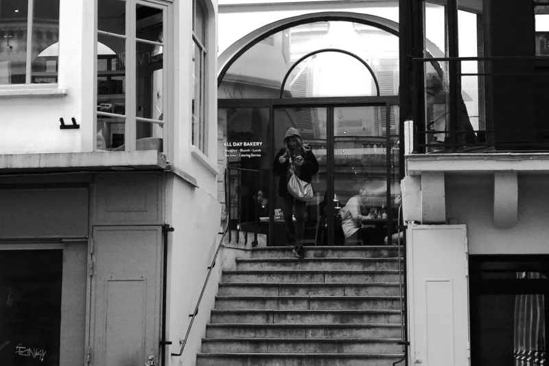 Bao · 30/10/2025 · 5472 x 3648 Cette photographie en noir et blanc capture un escalier extérieur menant à une entrée architecturale élégante. Le sujet principal semble être une scène urbaine avec des personnes assises sur les marches, dont une semble jouer de la musique.La composition repose sur une perspective centrale, avec un cadrage serré sur l’escalier et l’arche en fond. La lumière, douce et diffuse, accentue les contrastes sans être trop forte. L’ambiance est calme et mélancolique, renforcée par le monochrome. Le style rappelle le photojournalisme classique, évoquant une atmosphère intemporelle et contemplative.