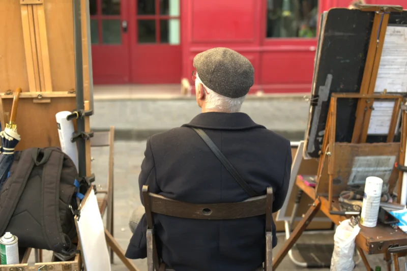 Bao · 6024 x 4016 Cette photographie montre un homme, sujet principal, vu de dos, assis et peinturant devant une toile sur un chevalet. La composition est simple, centrée sur l’artiste en plein travail. Le cadrage est serré, mettant en avant ses actions et son environnement immédiat.La lumière semble naturelle, douce, éclairant la scène sans créer d’ombres marquées. Les couleurs dominantes sont sobres : tons neutres (noir de la veste, gris du sol) et touches vives (rouge du bâtiment en arrière-plan).Le style est réaliste et documentaire, capturant un moment authentique. L’ambiance est calme et concentrée, évoquant la sérénité de la création artistique.