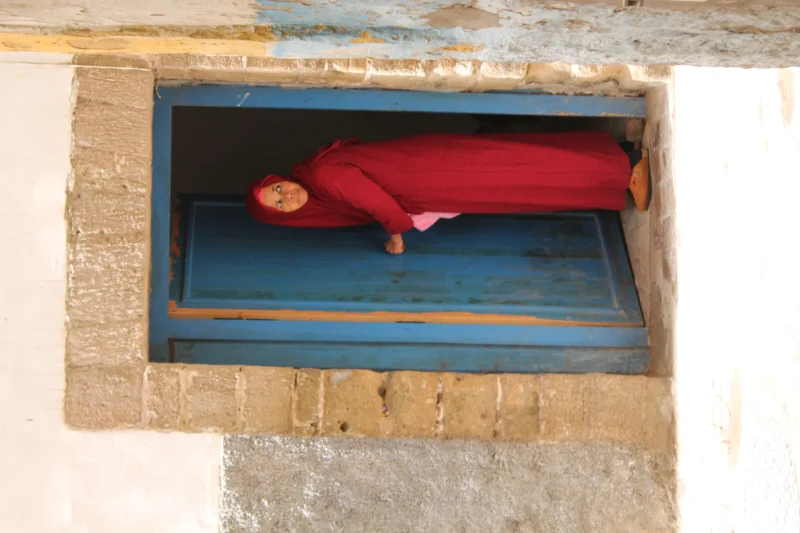 Sophie · 06/09/2025 · 4272 x 2848 Cette photographie montre un enfant en robe rouge allongé dans un petit espace rectangulaire encadré, probablement une fenêtre ou un passage étroit. La composition est simple et centrée, avec un cadrage serré qui met en valeur le sujet.La lumière semble naturelle, douce et légèrement tamisée, créant des ombres douces. Les couleurs dominantes sont le rouge vif de la robe et le bleu profond de l'encadrement. Le style est réaliste et documentaire, évoquant une ambiance à la fois poétique et mystérieuse, presque onirique.