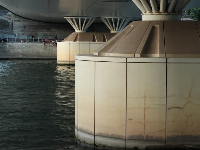 Francoise · 23/05/2026 · 6000 x 4000 Cette photographie met en scène un **pilier architectural** partiellement immergé dans l’eau, probablement un élément d’un pont ou d’un quai. La **composition** est épurée, avec une perspective basse accentuant la verticalité du sujet. Le **cadrage** serré isole ce pilier, tandis que l’arrière-plan flou et les structures métalliques ajoutent de la profondeur.La **lumière** est douce et naturelle, créant des contrastes subtils entre ombres et reflets. Les **couleurs dominantes** sont des tons neutres (beige, gris) et des reflets bleutés de l’eau, renforçant une ambiance **minimaliste et industrielle**. Le style évoque une **esthétique contemporaine**, avec une touche mélancolique et apaisante.