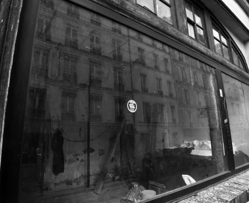 Bao · 2945 x 2402 Cette photographie en noir et blanc montre une scène reflétée dans une vitrine. Le sujet principal est une rue délabrée avec des bâtiments endommagés et une voiture abandonnée. La composition joue sur la réflexion et le cadrage serré, capturant des détails réalistes.La lumière est tamisée, créant des contrastes marqués. Les couleurs dominantes, bien que limitées au noir et blanc, suggèrent des tons grisâtres et sombres. Le style est documentaire et réaliste, évoquant une ambiance mélancolique et post-apocalyptique, renforcée par le désordre et l'abandon visible.