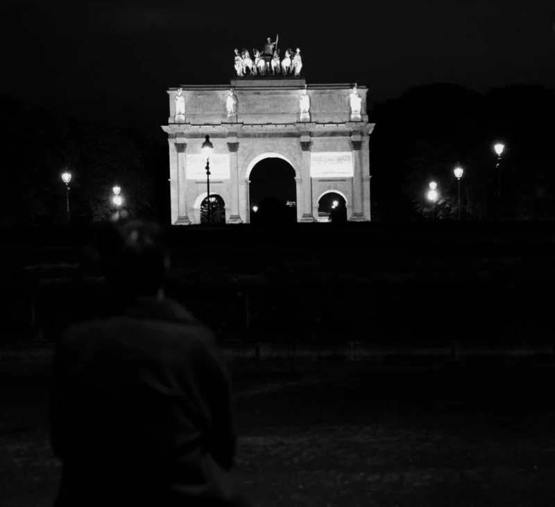 Bao · 6535 x 5963 Cette photographie en noir et blanc met en scène l'Arc de Triomphe de Munich, sujet principal, éclairé la nuit. La composition est symétrique, avec l'arc au centre, encadré par des arbres sombres. Le cadrage est large, incluant une partie du premier plan où une silhouette floue suggère la présence d'une personne.Les jeux de lumière contrastent fortement entre l'arc illuminé et l'obscurité environnante. Les couleurs dominantes sont les noirs et blancs, renforçant l'atmosphère dramatique. Le style est photographique classique, avec une ambiance nocturne mystérieuse et solennelle.