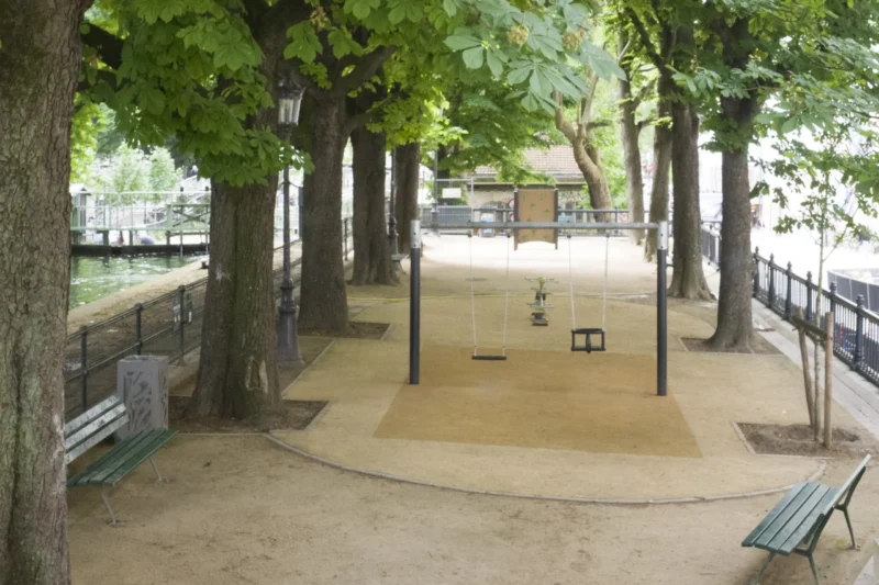 Dominique · 20/05/2026 · 2955 x 1970 Cette photographie montre un espace extérieur ombragé, probablement un ancien manège ou une aire de jeux.**Sujet principal** : Une piste circulaire en sable entourée de structures en bois.
**Composition** : Symétrique, avec des arbres en bordure et des bancs de chaque côté.
**Cadrage** : Vue large, légèrement en plongée.
**Lumière** : Naturelle et tamisée, filtrée par les feuilles.
**Couleurs dominantes** : Verts (feuillage), gris-beige (sable et bois), touches de brun.
**Style** : Documentaire, sobre et épuré.
**Ambiance** : Calme et nostalgique, évoquant un lieu peu fréquenté.