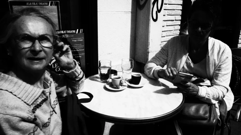 Dominique · 20/05/2026 · 6192 x 3480 Cette photographie en noir et blanc montre deux hommes âgés dans un café. Le sujet principal semble être l'interaction entre eux : l'un parle au téléphone, l'autre lit ou écrit.La composition est symétrique avec une table centrale. Le cadrage est serré, mettant en valeur les visages et les mains. La lumière est douce et naturelle, éclairant les sujets sans ombres marquées. L'ambiance est intime et calme, évoquant une scène du quotidien. Le style est documentaire, réaliste, avec une touche de mélancolie. Les tons clairs et foncés renforcent l'atmosphère rétro et authentique.