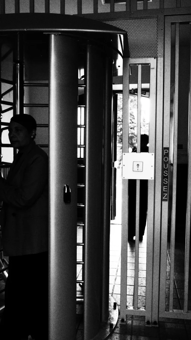 Dominique · 20/05/2026 · 3124 x 5558 Cette photographie en noir et blanc montre un sujet principal flou, probablement une personne à l’intérieur d’un espace éclairé, comme un bâtiment ou un véhicule. La composition met en avant un contraste entre l’obscurité extérieure et la lumière intérieure, créant un jeu de clarté et d’ombre.Le cadrage est serré, centré sur une porte ou une ouverture, avec une silhouette partielle à gauche. La lumière est douce mais contrastée, accentuant les contours. Les couleurs dominantes sont les tons de gris, renforçant l’ambiance mystérieuse et dramatique. Le style rappelle le photojournalisme ou un instantané documentaire, évoquant une atmosphère nocturne et intimiste.