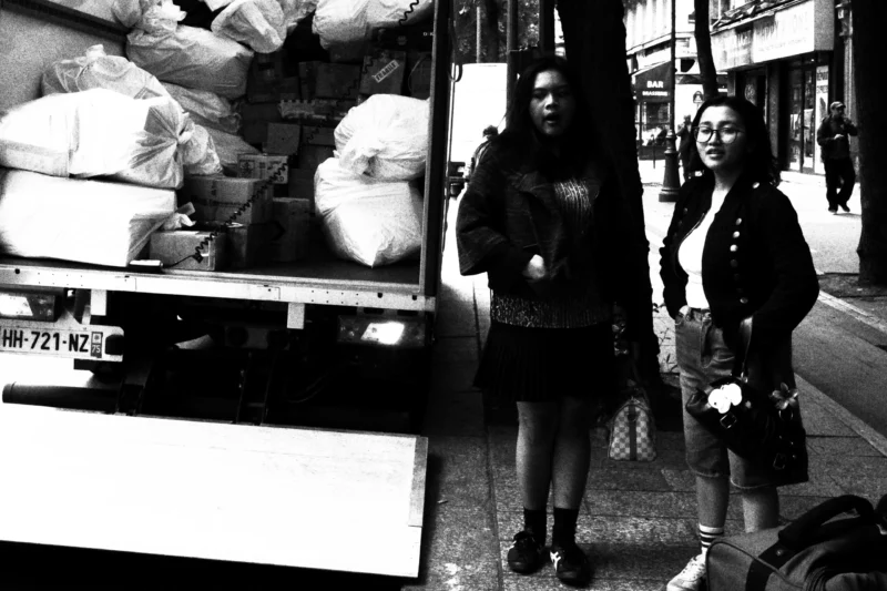 Dominique · 20/05/2026 · 3724 x 2482 Cette photographie en noir et blanc capture deux jeunes femmes comme sujets principaux, marchant sur un trottoir. La composition montre une rue urbaine avec des sacs empilés sur un camion en arrière-plan.Le cadrage est serré sur les deux femmes, avec une profondeur de champ qui met l'accent sur elles. La lumière est naturelle, probablement en journée, mais contrastée, créant des ombres marquées. Les couleurs dominantes sont les nuances de gris, typiques du noir et blanc.Le style est documentaire et spontané, évoquant une ambiance authentique et quotidienne des années 1970 ou 1980.