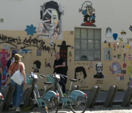 Bao · 6024 x 3452 Cette photographie montre un groupe de personnes devant un mur tagué et coloré. Le **sujet principal** est le mur recouvert de graffitis, portraits et symboles, notamment des étoiles de David.La **composition** est dynamique, avec des bancs et des vélos en premier plan. Le **cadrage** est large, incluant un espace urbain animé. La **lumière** semble naturelle, probablement en journée, avec des ombres douces.Les **couleurs dominantes** sont vives et contrastées : bleu, noir, blanc, rouge et jaune. Le **style** est street art, mêlant réalisme et symbolisme. L’**ambiance** est à la fois artistique et quotidienne, reflétant une scène de vie urbaine et culturelle.