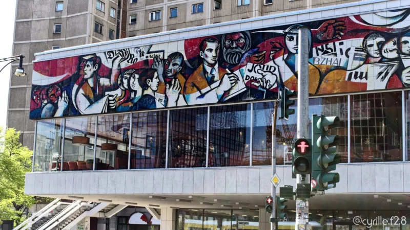 Cyrille · 30/04/2026 · 2026 x 1140 Cette photographie montre un bâtiment urbain orné d’un **mural coloré** représentant des personnages stylisés. Le **sujet principal** est ce graffiti dynamique, occupant presque toute la façade.La **composition** est équilibrée, avec un **cadrage serré** sur le mur, encadré par des éléments architecturaux modernes. La **lumière** semble naturelle, bien que légèrement contrastée, mettant en valeur les détails des couleurs vives.Les **couleurs dominantes** sont des tons **vifs** (rouge, bleu, jaune) et des contrastes noirs et blancs. Le **style** est graphique et expressif, proche du street art. L’**ambiance** est urbaine et artistique, mêlant modernité et créativité.