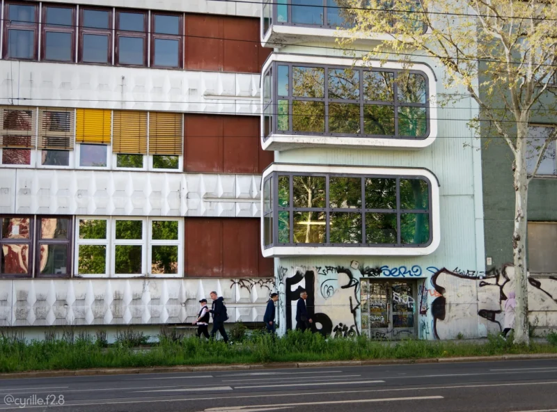Cyrille · 30/04/2026 · 2026 x 1498 Cette photographie montre un bâtiment moderne avec des extensions vitrées en saillie, abritant probablement des espaces végétalisés ou des serres urbaines. Le sujet principal est cette structure architecturale innovante.La composition met en valeur la juxtaposition entre l’ancien (bâtiment en brique) et le moderne (extensions en verre). Le cadrage inclut une partie de la rue avec des cyclistes, ajoutant une touche de vie quotidienne.La lumière est naturelle, douce, et semble provenir d’un ciel légèrement nuageux. Les couleurs dominantes sont les tons gris, verts et bruns, avec des touches de blanc et de bleu. Le style est contemporain et urbain, créant une ambiance à la fois futuriste et intégrée dans un environnement réel.