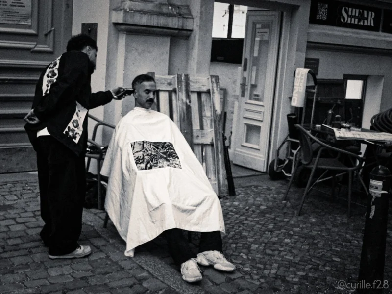 Cyrille · 30/04/2026 · 2026 x 1520 Cette photographie en noir et blanc montre un barbu assis sous une cape de coiffure, recevant une coupe de cheveux dans la rue. Le sujet principal est le client et le coiffeur en action.La composition est dynamique, avec un cadrage serré sur les deux personnages. L’arrière-plan, légèrement flou, met en valeur un cadre urbain avec des bâtiments et des tables de café. La lumière est naturelle, douce mais contrastée, accentuant les textures et les ombres. L’ambiance est réaliste et quotidienne, évoquant une scène de rue authentique et spontanée.