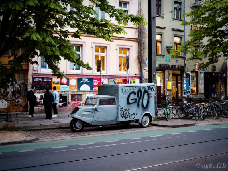 Cyrille · 30/04/2026 · 2026 x 1519 Cette photographie met en scène un petit utilitaire blanc marqué "G10" garé sur un trottoir urbain. La composition montre une rue animée avec des passants et des vélos en arrière-plan. Le cadrage est serré sur le véhicule, encadré par des arbres et des bâtiments.La lumière est douce et tamisée, typique d’un jour nuageux ou du soir. Les couleurs dominantes sont des tons neutres (gris, blanc) avec des éclats de couleurs vives sur les affiches et enseignes. Le style est réaliste et documentaire, capturant une scène de vie urbaine. L’ambiance est calme et légèrement mélancolique.