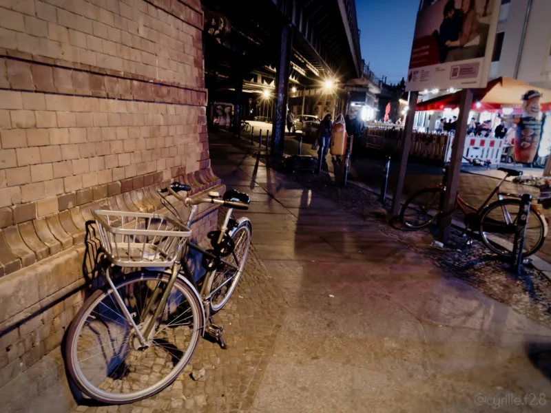 Cyrille · 30/04/2026 · 2026 x 1520 Cette photographie montre une ruelle nocturne avec des vélos comme sujet principal. La composition met en avant un vélo en premier plan, partiellement éclairé, tandis que d'autres vélos et des bâtiments sont visibles en arrière-plan.Le cadrage est serré sur les vélos et la ruelle étroite. La lumière est tamisée, avec des éclairages artificiels doux provenant des enseignes et des lampadaires, créant une ambiance chaleureuse et intimiste. Les couleurs dominantes sont des tons sombres et neutres, avec des éclats de bleu et de rouge.Le style est réaliste et documentaire, évoquant une scène de vie urbaine nocturne paisible.