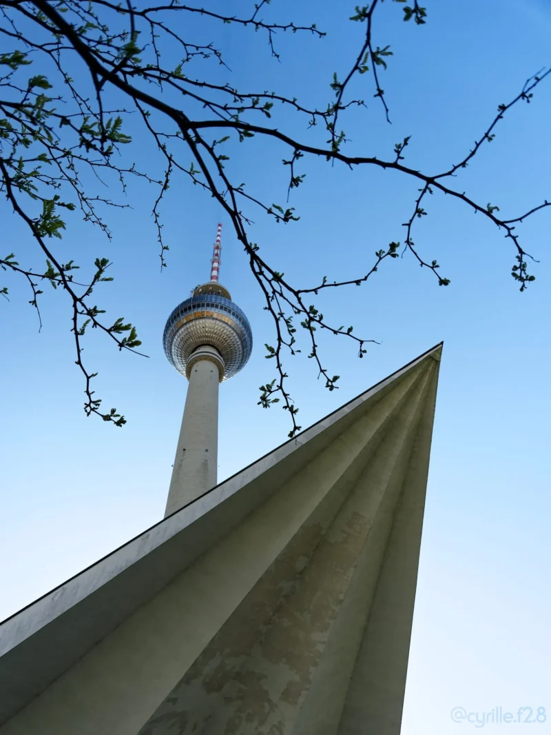Cyrille · 01/05/2026 · 1520 x 2026 Cette photographie met en scène la **Tour de télévision de Berlin** (Fernsehturm) en arrière-plan, partiellement cachée par des branches d’arbre en fleurs.La **composition** joue sur le **cadrage serré** d’un élément architectural en premier plan, probablement une pointe de toit ou une structure triangulaire, créant une perspective dynamique. La **lumière** est naturelle, douce et légèrement voilée, suggérant un jour clair mais pas ensoleillé. Les **couleurs dominantes** sont des tons pastel (blanc, bleu pâle) et des touches de vert clair des feuilles.Le style est **minimaliste et épuré**, évoquant une **ambiance calme et contemplative**, presque poétique.