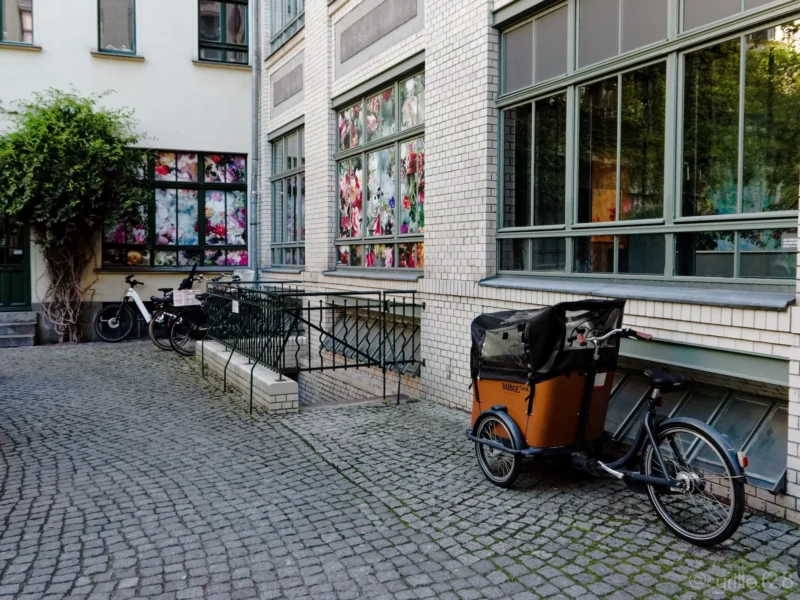 Cyrille · 01/05/2026 · 2026 x 1520 Cette photographie montre un **vélo-cargo** garé devant une boutique. Le sujet principal est ce vélo, avec sa remorque en bois et sa roue avant visible.La **composition** est simple et équilibrée, avec une perspective légèrement en contre-plongée. Le **cadrage** est serré sur le vélo et la façade du magasin, incluant une partie de la rue pavée.La **lumière** est naturelle et douce, probablement en journée. Les **couleurs dominantes** sont neutres (beige, gris) avec des touches de vert et de bois naturel. L’ambiance est **urbaine et paisible**, évoquant un quartier résidentiel ou commercial calme.