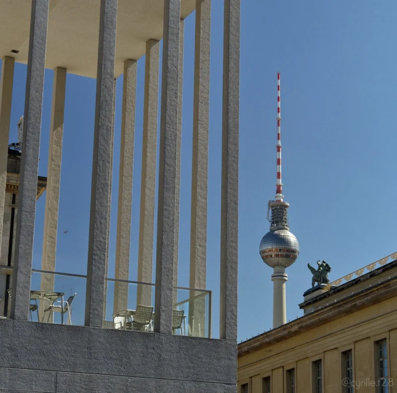 Cyrille · 01/05/2026 · 2026 x 2005 Cette photographie met en scène le **Fernsehturm de Berlin**, tour emblématique avec sa sphère métallique et son antenne rouge et blanche. Le cadrage est serré, intégrant aussi une partie d’un bâtiment moderne en premier plan avec ses colonnes verticales.La lumière est naturelle, éclatante, sous un ciel bleu pur. Les couleurs dominantes sont les **tons gris (bâtiment), bleu (ciel), et argenté (tour)**. Le style est documentaire et architectural, capturant une ambiance urbaine dynamique et moderne. L’image évoque une ville contrastée entre tradition et avant-garde.