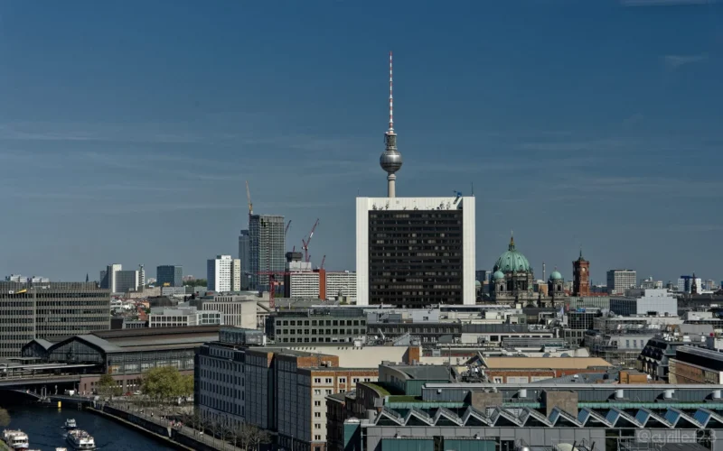Cyrille · 01/05/2026 · 2026 x 1266 Cette photographie montre la ville de Berlin avec pour sujet principal la **Fernsehturm**, tour de télévision emblématique. La composition met en valeur cette structure centrale, entourée de bâtiments variés. Le cadrage est large, offrant une vue panoramique sur l'horizon urbain.La lumière est naturelle, avec un ciel dégagé, créant des contrastes nets. Les couleurs dominantes sont les **tons gris et beiges** des bâtiments, adoucis par des touches de vert et de bleu. Le style est documentaire et réaliste, évoquant une ambiance moderne et dynamique, typique d'une métropole européenne.