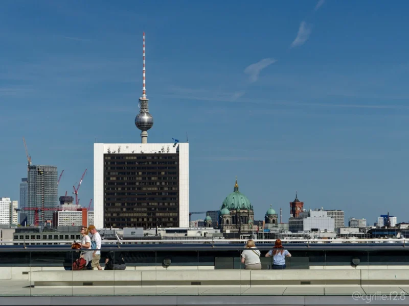 Cyrille · 01/05/2026 · 2026 x 1520 Cette photographie montre la **Tour de télévision de Berlin** (Fernsehturm) en sujet principal, avec sa structure distinctive et sa sphère dorée. La composition inclut un cadrage large, intégrant des bâtiments urbains et des éléments architecturaux variés en arrière-plan.La lumière est naturelle, avec un ciel bleu clair, créant une ambiance lumineuse et dégagée. Les couleurs dominantes sont les **tons gris et blancs** des bâtiments, contrastés par le **bleu du ciel** et le **doré de la sphère**.Le style est photographique réaliste, avec une ambiance urbaine moderne et dynamique.