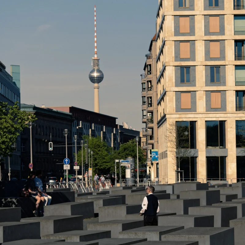 Cyrille · 01/05/2026 · 2026 x 2026 Cette photographie montre le **Fernsehturm de Berlin**, tour emblématique, en arrière-plan. La composition met en valeur une **place avec des blocs de béton mémoriels** (Mémorial aux Juifs assassinés d'Europe) et des bâtiments modernes.Le cadrage est large, incluant des éléments urbains et des passants. La lumière est naturelle, claire et douce, typique d’un jour ensoleillé. Les couleurs dominantes sont les **tons gris des bâtiments**, les **noirs des blocs mémoriels** et les **teintes bleutées du ciel**.Le style est **documentaire et urbain**, évoquant une ambiance à la fois **mémorielle et contemporaine**.