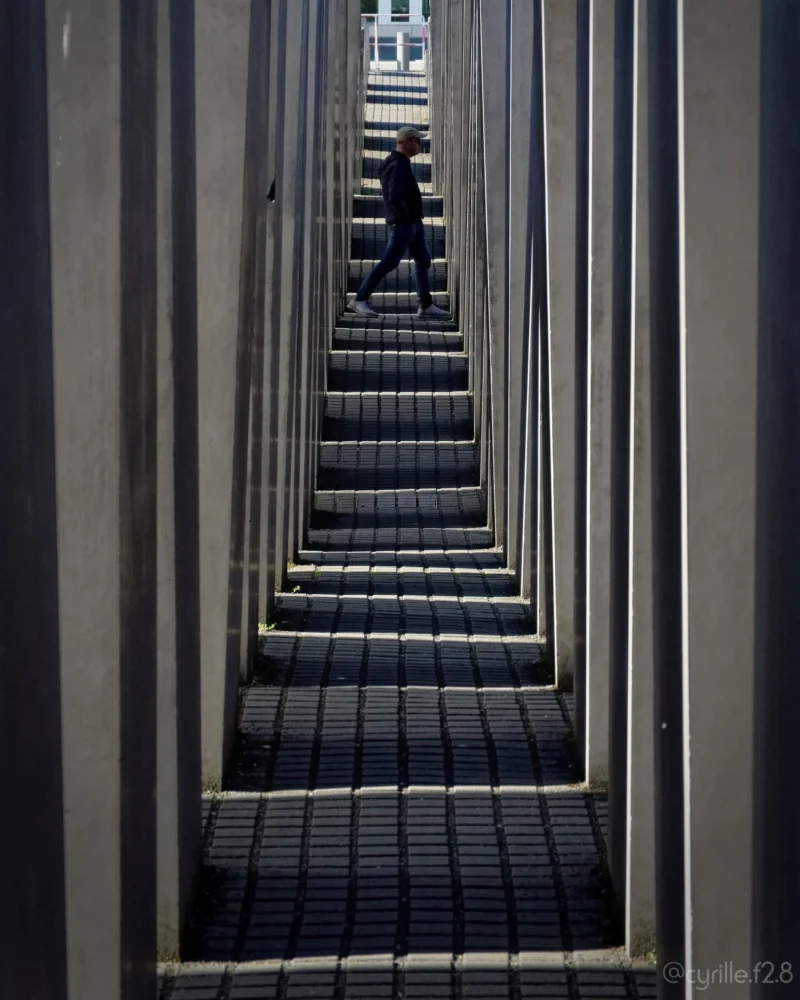 Cyrille · 01/05/2026 · 1621 x 2026 Cette photographie montre un escalier descendant en perspective répétée, créant un effet de tunnel. Le sujet principal est une personne en mouvement, presque floue, au centre des marches. La composition repose sur des lignes parallèles convergentes, renforçant la profondeur.Le cadrage est serré, avec une lumière tamisée et contrastée, mettant en valeur les ombres et les contours nets. Les couleurs dominantes sont les tons gris et bleus, avec des nuances sombres. Le style est épuré et géométrique, évoquant une ambiance à la fois mystérieuse et minimaliste, presque cinématographique.