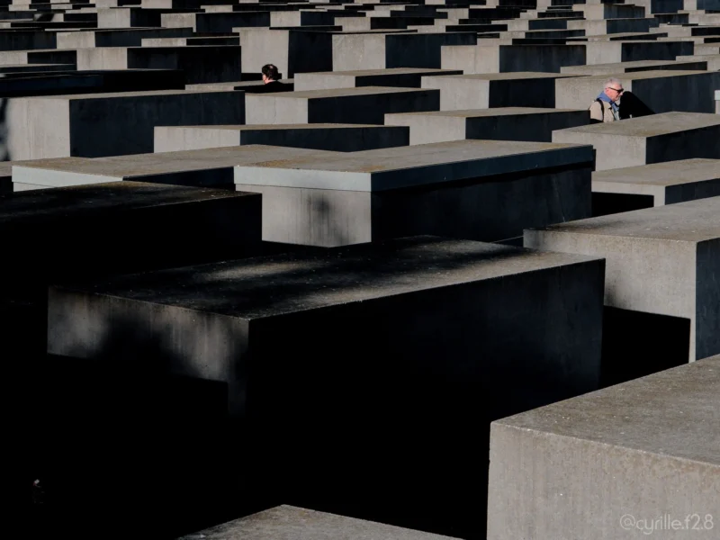 Cyrille · 01/05/2026 · 2026 x 1520 Cette photographie montre le **Mémorial aux Juifs assassinés d'Europe** à Berlin. Le **sujet principal** est l'immensité des **stèles rectangulaires en béton**, disposées en un labyrinthe uniforme.La **composition** est géométrique et répétitive, créant une ambiance oppressante. Le **cadrage** est serré, mettant en valeur les lignes parallèles et l'horizon lointain. La **lumière** est naturelle, douce et diffuse, adoucissant les contrastes.Les **couleurs dominantes** sont les gris et noirs, renforçant la gravité du lieu. Le **style** est épuré et minimaliste, typique de l'art mémoriel contemporain. L’**ambiance** est solennelle, presque mélancolique, invitant à la réflexion.