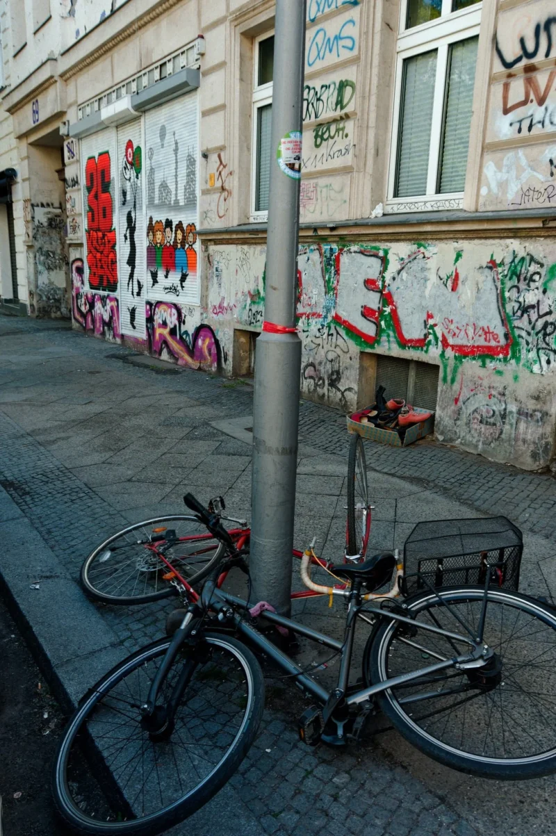 Cyrille · 02/05/2026 · 1347 x 2026 Cette photographie montre un vélo rouge et noir attaché à un poteau dans une rue urbaine. Le sujet principal est le vélo, mis en valeur par un cadrage serré.La composition inclut un mur couvert de graffitis colorés en arrière-plan, ajoutant une touche artistique. La lumière semble naturelle, avec des ombres douces, suggérant un éclairage matinal ou vespertin. Les couleurs dominantes sont les tons rouges, noirs, blancs et quelques touches de bleu et vert des graffitis.Le style est réaliste et documentaire, capturant une scène de vie urbaine. L’ambiance est à la fois dynamique et mélancolique, reflétant un mélange de créativité et de quotidien.