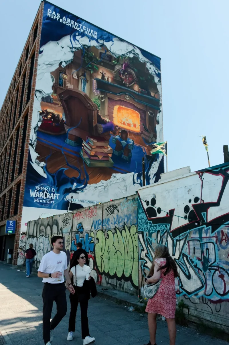 Cyrille · 02/05/2026 · 1347 x 2026 Cette photographie montre un groupe de personnes posant devant un grand mur coloré et graffité. Le sujet principal est le mural représentant une scène urbaine stylisée, avec des éléments de jeu de lumière et des personnages.La composition est dynamique, avec un cadrage serré sur le mural et les personnes en premier plan. La lumière est naturelle et éclatante, typique d’une journée ensoleillée. Les couleurs dominantes sont vives : bleu, orange, noir et blanc.Le style est urbain et artistique, mêlant street art et pop culture. L’ambiance est joyeuse et énergique, reflétant la créativité et la vie urbaine.