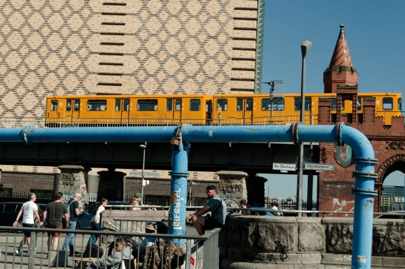 Cyrille · 02/05/2026 · 2026 x 1347 Cette photographie met en scène un **train jaune** en mouvement, sujet principal, traversant un pont métallique. La **composition** associe des éléments urbains et industriels : tuyaux bleus, rails, et une structure architecturale en arrière-plan.Le **cadrage** est serré sur le train et le pont, avec une perspective légèrement basse. La **lumière** est naturelle et vive, créant des contrastes nets. Les **couleurs dominantes** sont le jaune vif du train, le bleu des tuyaux et les tons gris et bruns des bâtiments.Le style est **documentaire et réaliste**, évoquant une ambiance **urbaine et dynamique**, mêlant modernité et infrastructure industrielle.