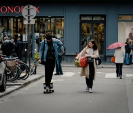 Cyrille · 16/05/2026 · 1200 x 900 Cette photographie capture une scène urbaine animée. Le sujet principal est une femme en mouvement sur un skateboard, vêtue d’une veste blanche et tenant un sac. La composition montre une rue étroite avec des passants en arrière-plan, dont une personne avec un parapluie.Le cadrage est serré sur la femme au premier plan, avec une profondeur de champ qui floute légèrement les éléments lointains. La lumière semble naturelle, probablement en journée, avec des ombres douces. Les couleurs dominantes sont neutres (gris, beige) avec des touches de rouge et de vert (parapluie).Le style est réaliste et dynamique, évoquant une ambiance moderne et quotidienne en ville.