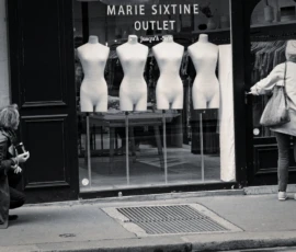 Cyrille · 16/05/2026 · 1200 x 798 Cette photographie en noir et blanc montre une scène urbaine devant une boutique "Marie Sixtine Outlet". Le sujet principal est une personne assise à gauche, probablement un sans-abri, contrastant avec une femme élégante à droite, qui semble faire ses courses.La composition oppose deux mondes sociaux. Le cadrage est serré, centré sur ces deux figures. La lumière est naturelle, avec des contrastes marqués. Les couleurs dominantes, en noir et blanc, accentuent les ombres et les textures.Le style est documentaire, réaliste, évoquant une ambiance sociale et contrastée, presque mélancolique.