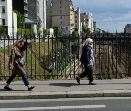 Cyrille · 16/05/2026 · 1200 x 900 Cette photographie capture deux personnes traversant un passage piéton en ville. Le sujet principal est ces deux individus, l’un vêtu d’un manteau clair et l’autre d’un costume sombre avec chapeau.La composition est symétrique, avec un cadrage serré sur le passage clouté. L’arrière-plan montre des immeubles et une clôture métallique. La lumière est naturelle et douce, typique d’une journée ensoleillée. Les couleurs dominantes sont les tons neutres (gris, beige) avec des touches de vert et bleu.Le style est documentaire et réaliste, évoquant une ambiance urbaine paisible et quotidienne.