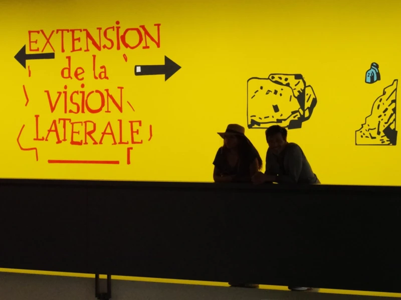 Agnes · 23/05/2026 · 5763 x 2819 Cette photographie montre deux silhouettes dos à la caméra devant un mur jaune vif. Le sujet principal est un message écrit en rouge : *« Extension de la vision latérale »*, avec des flèches et des dessins schématiques évoquant une perception élargie.La composition est épurée, centrée sur le texte et les silhouettes. Le cadrage est serré, mettant en valeur le contraste entre les formes humaines et le fond coloré. La lumière est naturelle et uniforme, éclairant clairement la scène. Les couleurs dominantes sont le jaune éclatant et le rouge vif, créant une ambiance dynamique et moderne, presque abstraite. Le style semble artistique et conceptuel.