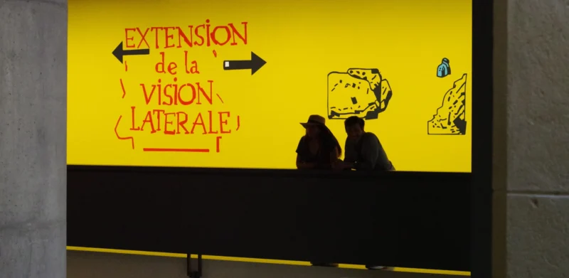 Agnes · 23/05/2026 · 5763 x 2819 Cette photographie montre deux silhouettes dos à la caméra devant un mur jaune vif. Le sujet principal est un message écrit en rouge : *« Extension de la vision latérale »*, avec des flèches et des dessins schématiques évoquant une perception élargie.La composition est épurée, centrée sur le texte et les silhouettes. Le cadrage est serré, mettant en valeur le contraste entre les formes humaines et le fond coloré. La lumière est naturelle et uniforme, éclairant clairement la scène. Les couleurs dominantes sont le jaune éclatant et le rouge vif, créant une ambiance dynamique et moderne, presque abstraite. Le style semble artistique et conceptuel.