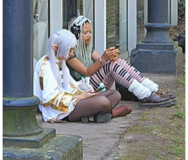 Sophie · 932 x 978 Cette photographie montre deux personnes assises, vêtues de costumes traditionnels colorés évoquant un style historique ou folklorique. Le sujet principal semble être leur interaction, l’une tenant un objet en forme de chaton.La composition est simple, avec un cadrage serré centré sur les deux personnages. L’arrière-plan, flou, met en valeur des colonnes et une végétation discrète. La lumière est naturelle, douce, créant des ombres légères. Les couleurs dominantes sont vives : or, blanc, noir et rouge, apportant une touche chaleureuse et festive.L’ambiance est à la fois intimiste et théâtrale, évoquant une scène de rue ou une représentation culturelle.