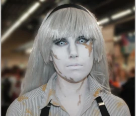 Sophie · 24/05/2026 · 1643 x 1531 Cette photographie met en scène un mannequin ou une personne maquillée de façon spectaculaire, évoquant un style androgyne et gothique. Le sujet principal, aux cheveux blonds platine et au teint pâle, arbore un maquillage exagéré avec des traits marqués et des ombres bleutées.Le cadrage est serré, centré sur le visage. La lumière semble artificielle, mettant en valeur les contrastes. Les couleurs dominantes sont le blanc, le bleu pâle et les tons grisâtres, renforçant une ambiance froide et mystérieuse. L’arrière-plan flou suggère un lieu public, peut-être une convention ou un événement artistique. L’ensemble dégage une atmosphère théâtrale et avant-gardiste.