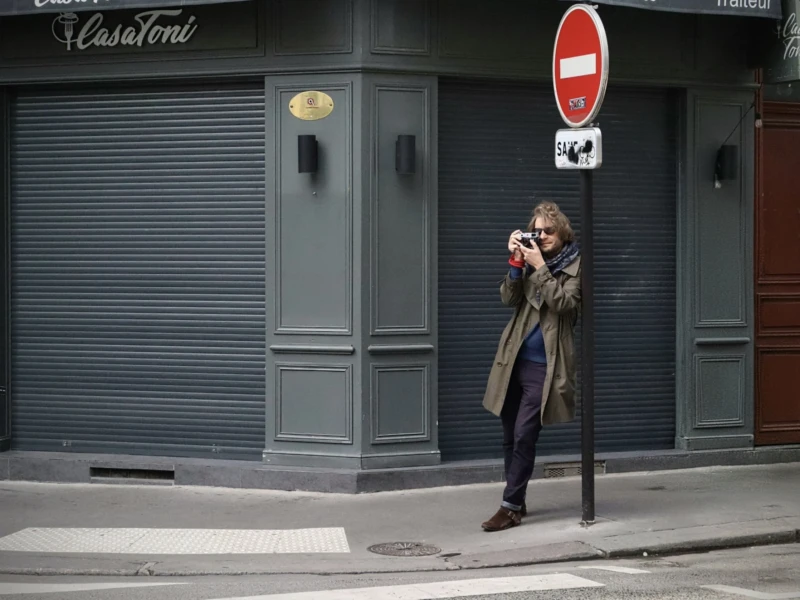 Elisa Ma · 16/05/2026 · 5596 x 3910 Cette photographie met en scène une personne comme sujet principal, prise en train de photographier un bâtiment. La composition est simple et épurée, avec un cadrage serré sur le sujet et l’arrière-plan minimaliste.La lumière semble naturelle, probablement en journée, mais un peu tamisée, créant des ombres douces. Les couleurs dominantes sont les tons gris (bâtiment, vêtements) et marron, avec une touche de rouge vif (panneau d’interdiction).Le style est réaliste et documentaire, évoquant une ambiance urbaine et calme, presque intimiste.