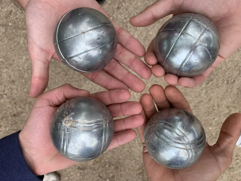Kerry · 03/05/2026 · 4032 x 3024 Cette photographie montre quatre boules en métal, probablement en laiton, tenues dans des mains. Le sujet principal est ces boules de pétanque, disposées en carré.Le cadrage est serré, centré sur les mains et les boules. La lumière naturelle éclaire uniformément la scène, sans ombres marquées. Les couleurs dominantes sont les tons gris métalliques des boules et les teintes naturelles des mains et du sol.Le style est simple et direct, avec une ambiance réaliste et quotidienne. L'image évoque un moment informel, presque artisanal.