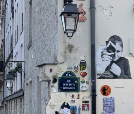 Kerry · 09/05/2026 · 3010 x 3923 Cette photographie montre une façade urbaine décorée de street art. Le sujet principal est un mélange d'éléments : une lanterne ancienne, des stickers variés (dont un Eiffel), des graffitis, et des affiches collées.La composition est verticale, avec un cadrage serré sur les détails. La lumière semble naturelle, douce, mettant en valeur les textures et les contrastes. Les couleurs dominantes sont neutres (gris, blanc) avec des touches vives (bleu, rouge, noir).Le style est urbain et éclectique, reflétant une ambiance artistique et vivante, typique des rues créatives.