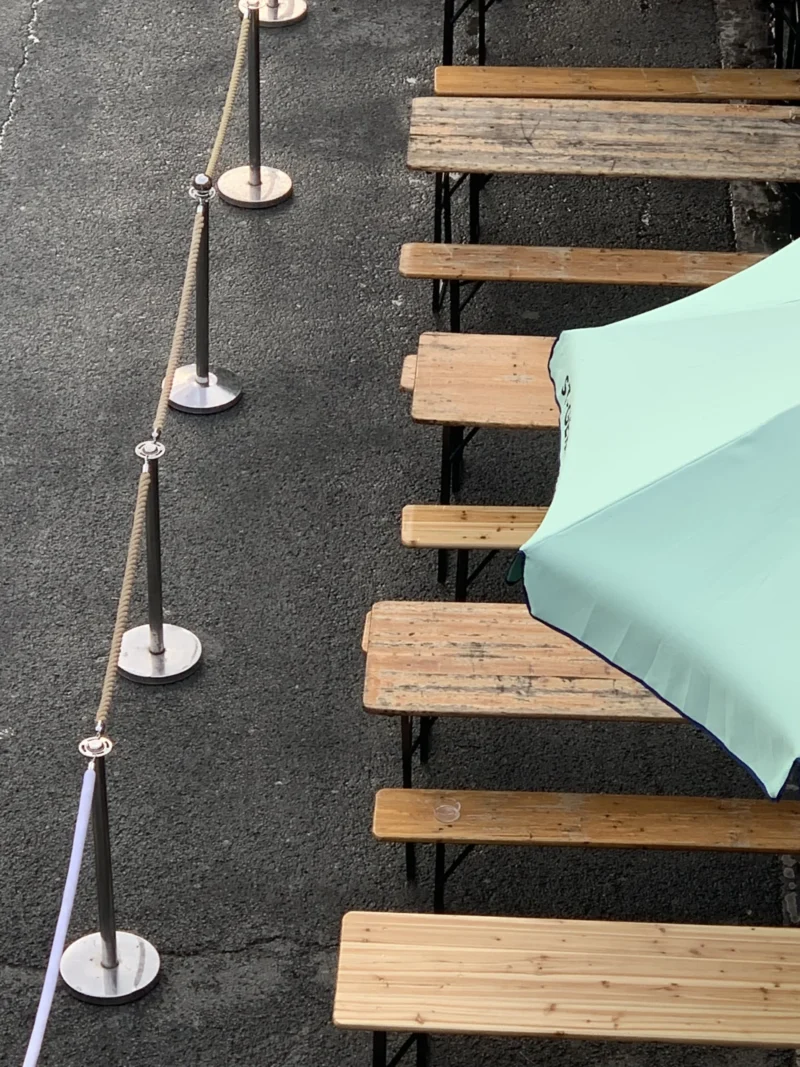 Kerry · 15/05/2026 · 4032 x 3024 Cette photographie montre des bancs en bois disposés sur une surface asphaltée, avec des barrières métalliques alignées à gauche. Le sujet principal est un parasol bleu pâle fermé, posé sur l’un des bancs.La composition est simple et géométrique, avec un cadrage serré sur les bancs et les barrières. La lumière semble naturelle, bien que légèrement diffuse, créant des ombres douces. Les couleurs dominantes sont les tons de bois naturel, le gris de l’asphalte et le bleu clair du parasol.Le style est épuré et minimaliste, évoquant une ambiance calme et ordonnée, typique d’un espace public ou d’une zone de détente urbaine.