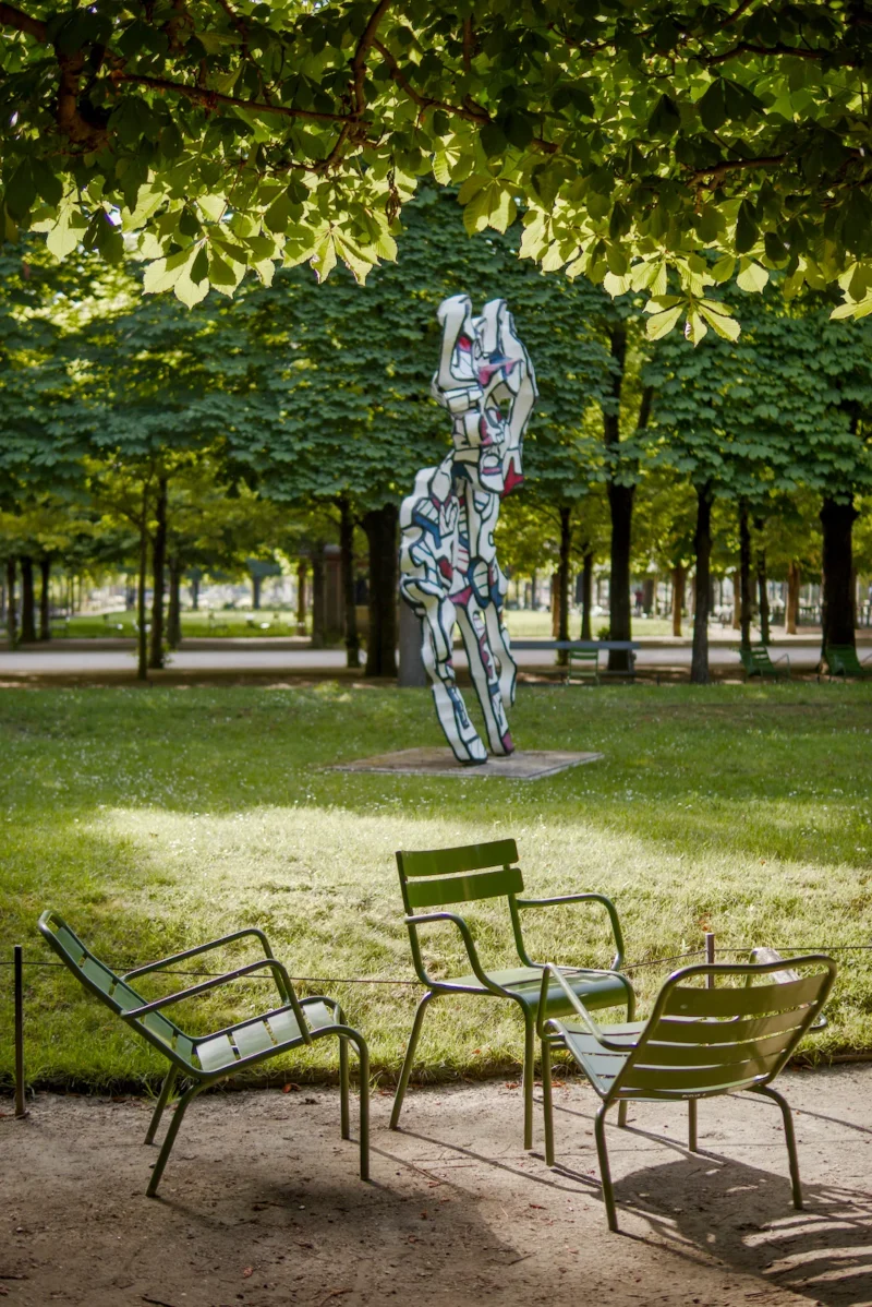 Frédéric · 24/05/2026 · 1121 x 1679 Cette photographie met en scène une **sculpture métallique abstraite** en forme de colonne fragmentée, entourée d’un **parc arboré**. La composition joue sur le contraste entre l’art contemporain et la nature, avec un **cadrage serré** sur les bancs vides en premier plan et l’œuvre en arrière-plan.La lumière est **naturelle et douce**, filtrée par les feuilles, créant des ombres légères. Les **couleurs dominantes** sont les verts profonds des arbres et les tons gris-bleutés de la sculpture. Le style est **épuré et minimaliste**, évoquant une ambiance **calme et contemplative**, presque onirique.
