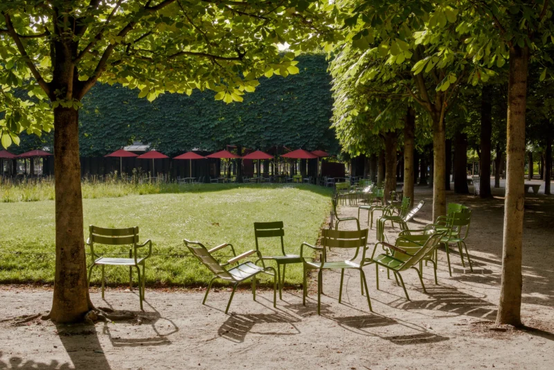 Frédéric · 24/05/2026 · 2517 x 1680 Cette photographie montre un espace extérieur paisible, probablement un parc ou une terrasse de café. Le sujet principal est une série de tables et chaises en métal disposées sur une allée en gravier, bordée d’arbres.La composition est symétrique et ordonnée, avec un cadrage en profondeur. La lumière, douce et naturelle, semble provenir des côtés, créant des ombres légères. Les couleurs dominantes sont les verts des arbres et du gazon, avec des touches de gris (gravier) et de rouge (parasols).Le style est épuré et minimaliste, évoquant une ambiance calme et invitante, presque nostalgique.
