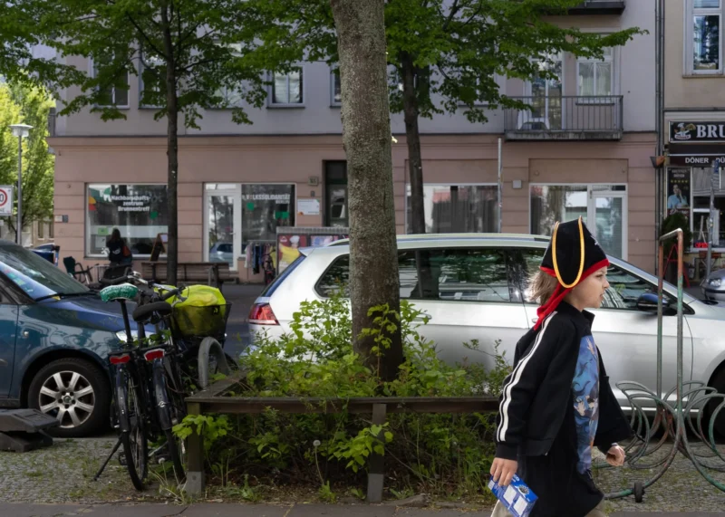 Muriel · 30/04/2026 · 2048 x 1463 Cette photographie met en scène une personne en mouvement, vêtue d’un manteau sombre et coiffée d’un chapeau jaune, comme sujet principal. La composition montre une rue urbaine avec des voitures, des bâtiments et des arbres en arrière-plan.Le cadrage est serré sur la personne, avec une profondeur de champ qui floute légèrement l’arrière-plan. La lumière est naturelle, probablement en journée, avec des ombres douces. Les couleurs dominantes sont les tons neutres (gris des bâtiments, vert des arbres) et les touches vives (jaune du chapeau, bleu du manteau).L’ambiance est calme et quotidienne, typique d’un environnement urbain paisible.