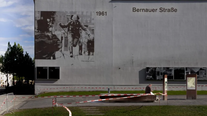 Muriel · 30/04/2026 · 2048 x 1152 Cette photographie montre un mur extérieur avec une grande fresque datant de 1961, située "Bernauer Straße". Le sujet principal est une illustration en noir et blanc représentant des personnes en situation difficile, évoquant un contexte historique lourd.La composition est simple, avec un cadrage serré sur le mur et un sol partiellement visible. La lumière est naturelle, douce mais contrastée, créant des ombres nettes. Les couleurs dominantes sont les tons gris et blancs, renforcés par la fresque monochrome. L’ambiance est sobre et mélancolique, évoquant la mémoire et l'histoire.