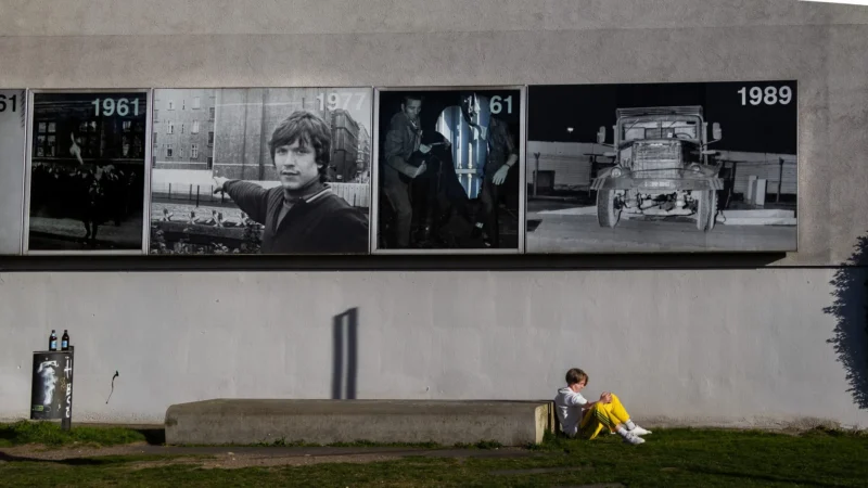 Muriel · 30/04/2026 · 2048 x 1152 Cette photographie montre une personne assise sur un banc devant un mur décoré de trois panneaux photographiques en noir et blanc, datés 1981, 1987 et 1989. Le sujet principal est cette personne, vêtue d’un manteau jaune, qui semble contemplative.La composition oppose les images historiques et contemporaines. Le cadrage est serré sur le banc et le mur, avec une lumière naturelle douce et diffuse. Les couleurs dominantes sont les tons gris des photos et le jaune vif du manteau, créant un contraste saisissant. L’ambiance est à la fois mélancolique et introspective, évoquant une réflexion sur le temps et la mémoire.