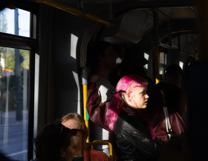 Muriel · 30/04/2026 · 2048 x 1583 Cette photographie montre une personne vêtue d’un costume coloré, évoquant un personnage de super-héros, assis dans un bus. Le sujet principal est flou mais reconnaissable grâce à sa tenue rose et noire.La composition met en valeur le contraste entre le personnage et les autres passagers peu visibles. Le cadrage est serré, centré sur lui, avec une lumière naturelle filtrant par les fenêtres, créant des ombres marquées. Les couleurs dominantes sont les tons roses, noirs et gris, renforçant une ambiance à la fois mystérieuse et dynamique. Le style est réaliste et spontané, capturant un moment du quotidien avec une touche originale.