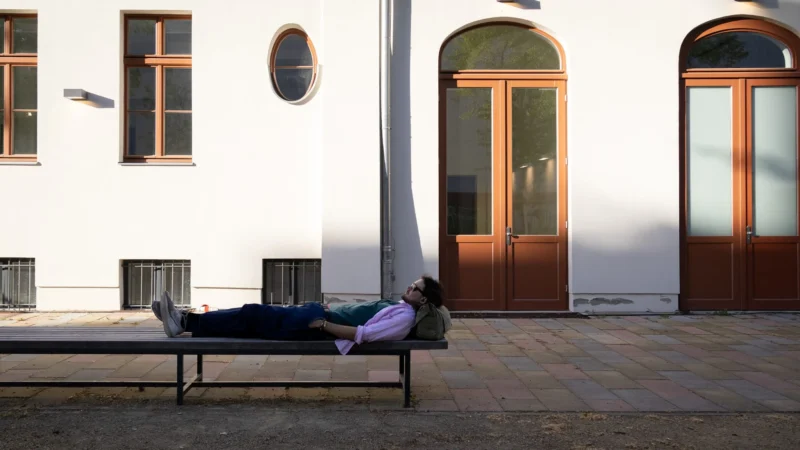 Muriel · 30/04/2026 · 2048 x 1152 Cette photographie montre un homme allongé sur un banc, dormant ou se reposant, devant un bâtiment blanc. Le sujet principal est cet homme, isolé dans un espace urbain calme.La composition est simple et épurée, avec un cadrage serré sur le banc et le personnage. La lumière naturelle, douce et légèrement dorée, suggère un moment en fin de journée. Les couleurs dominantes sont les tons blancs et beiges du bâtiment, contrastant avec le gris du sol et les teintes sombres des vêtements de l’homme.L’ambiance est paisible et mélancolique, évoquant la solitude et la tranquillité. Le style est réaliste et documentaire.